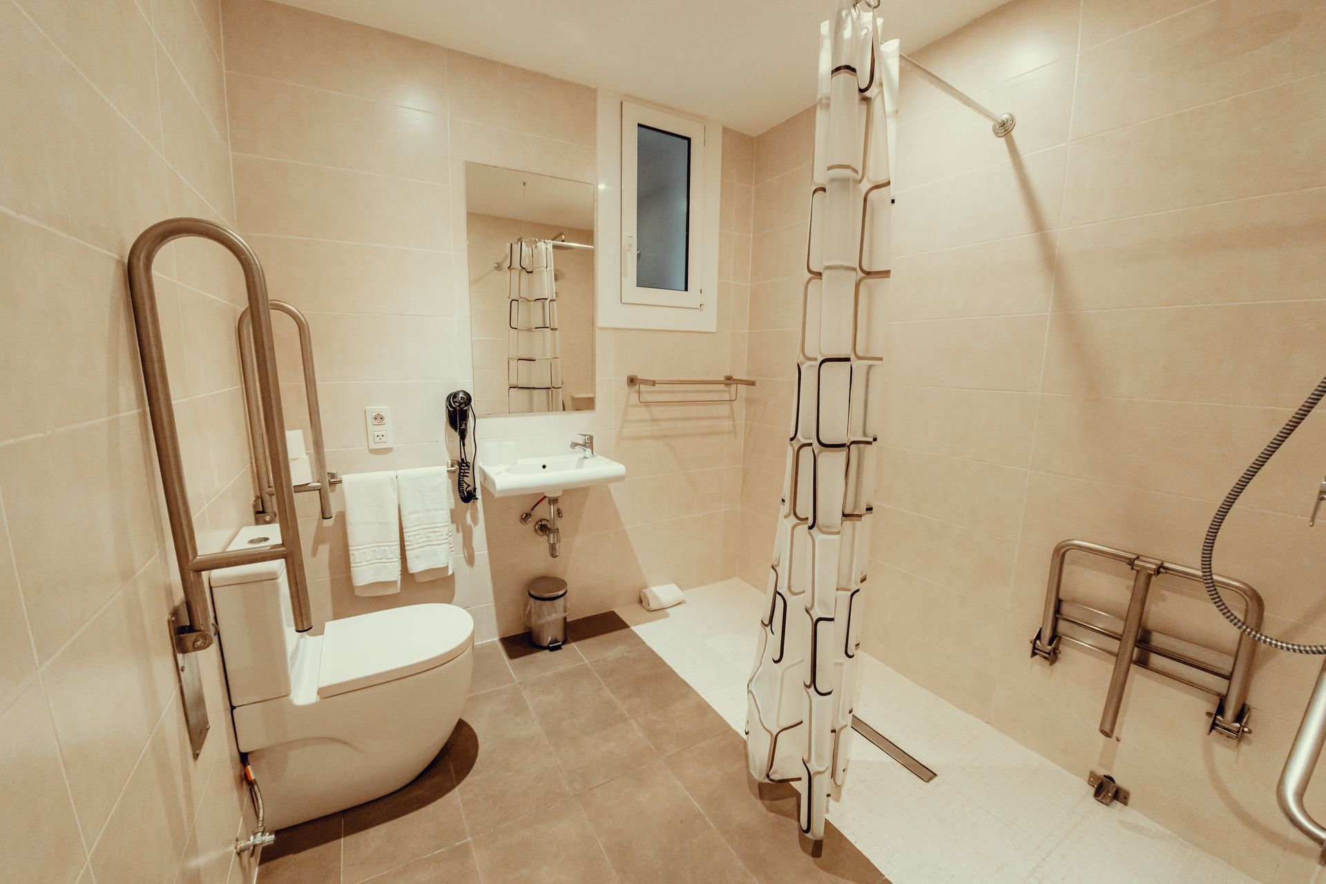 Baño accesible con inodoro, lavabo, silla de ducha, barras de apoyo y ducha a ras de suelo con azulejos de color beige.