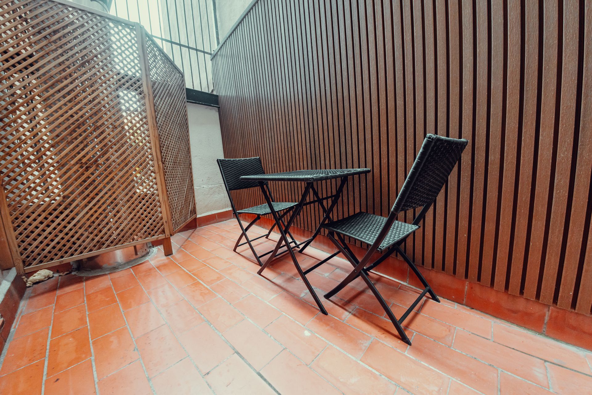 Patio exterior con dos sillas plegables negras y una mesita junto a una pared de madera y una celosía.