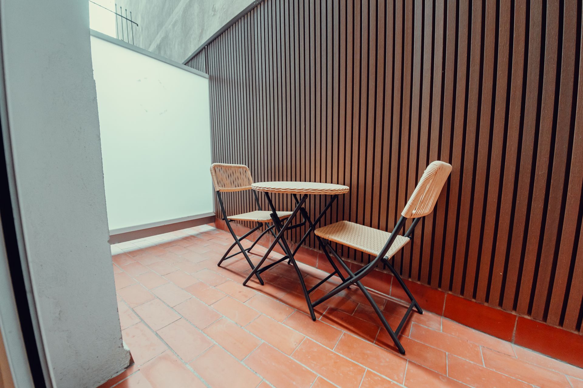 Pequeño balcón con dos sillas plegables y una mesa redonda junto a una pared de listones de madera.