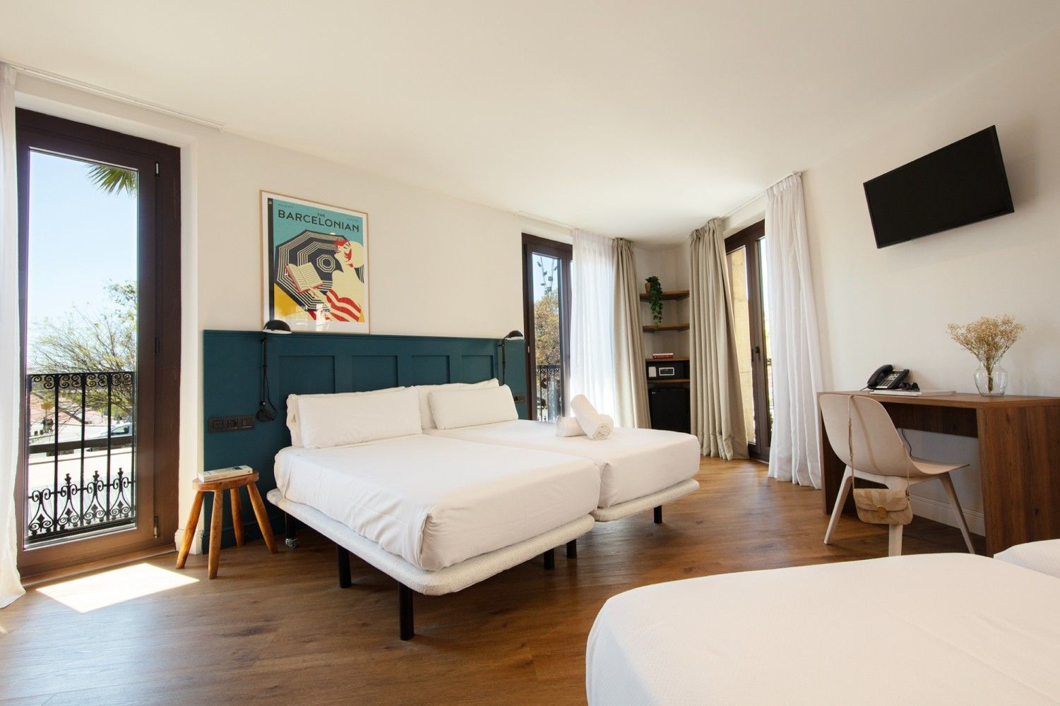 Una habitación de hotel con dos camas blancas sobre una cabecera verde azulado, un escritorio de madera con una silla.