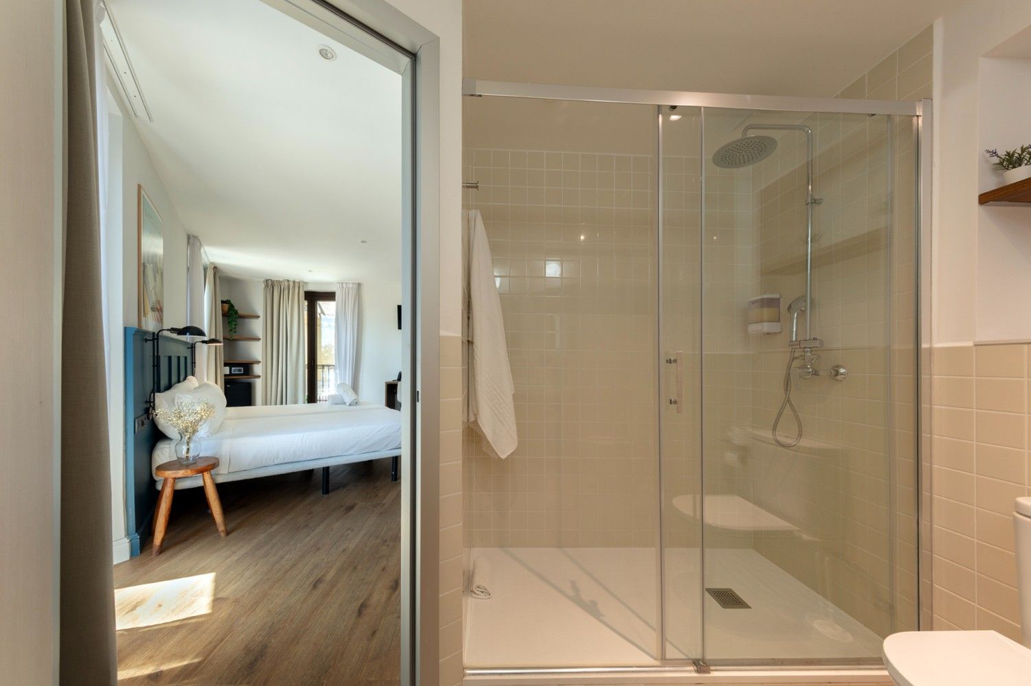 Una ducha con mampara de cristal en un baño moderno, con vista a través de una puerta a un dormitorio con una cama.