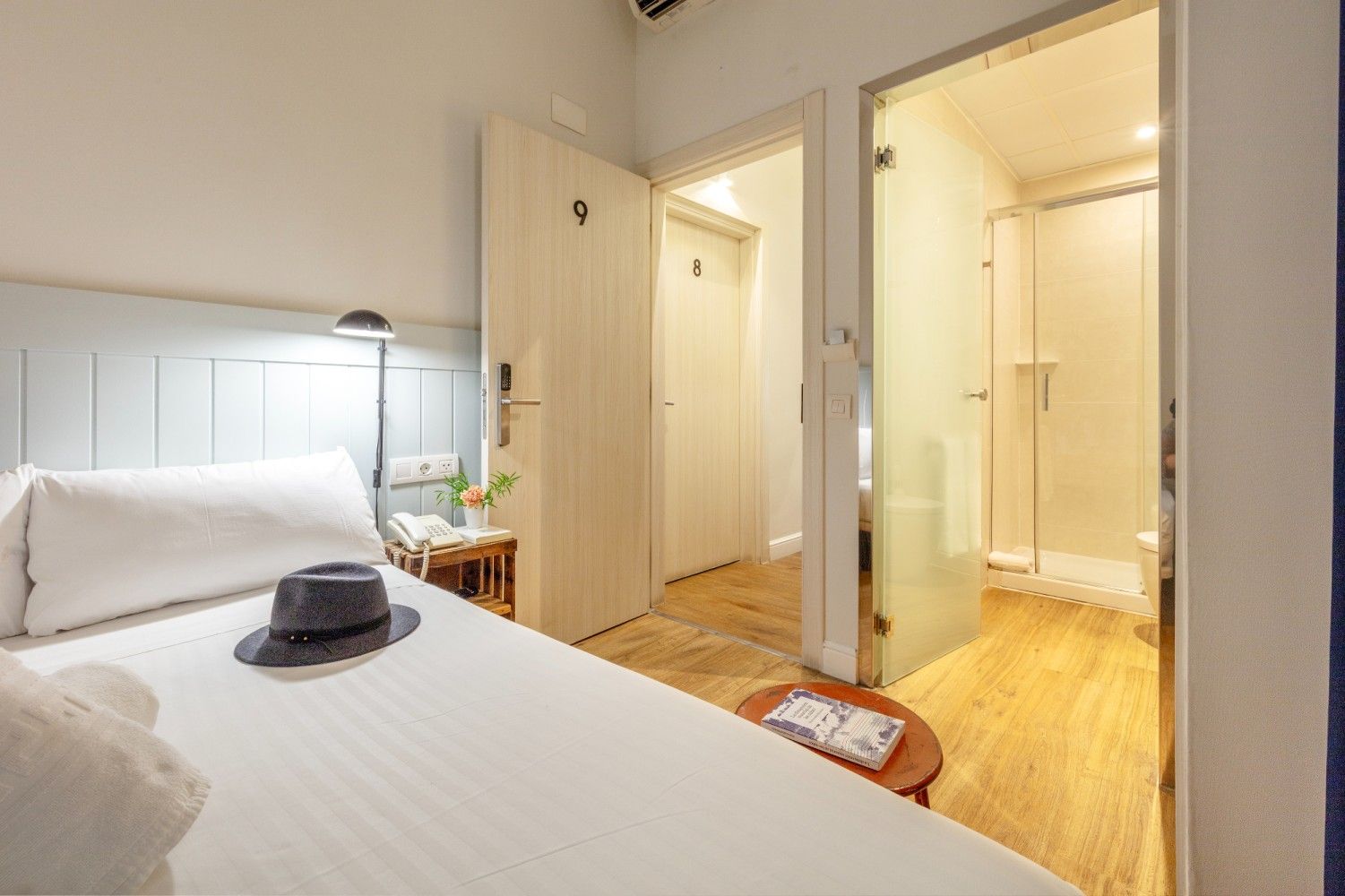 Una habitación de hotel con una cama blanca, un sombrero gris, una mesita de noche.