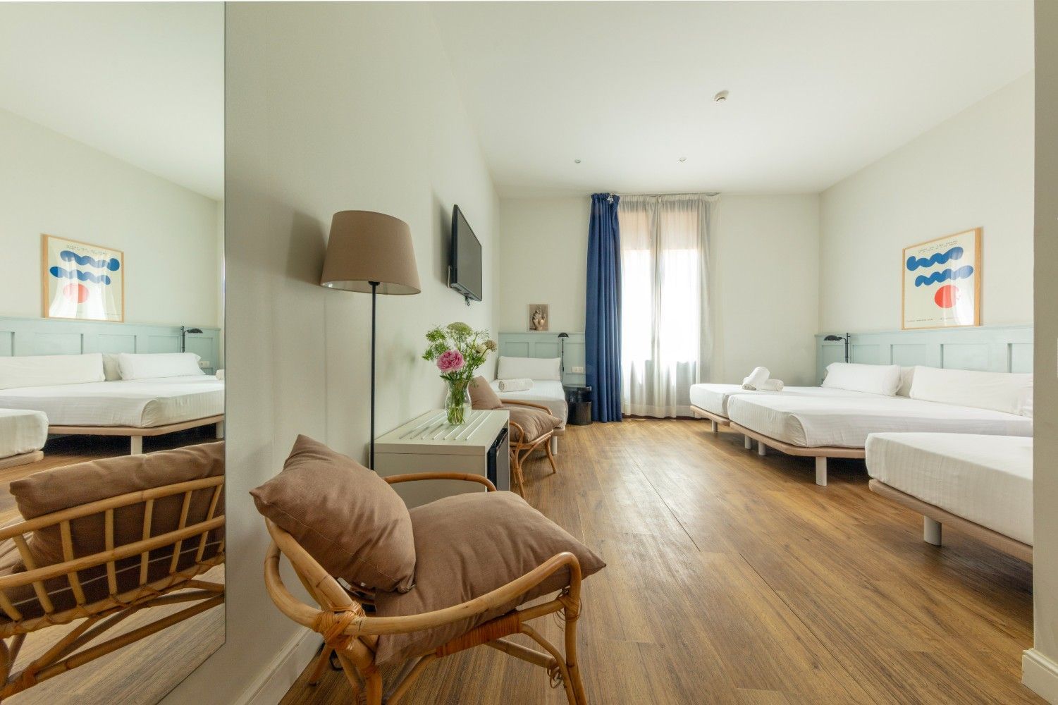 Una habitación de hotel luminosa con pisos de madera clara, varias camas, una silla de ratán.