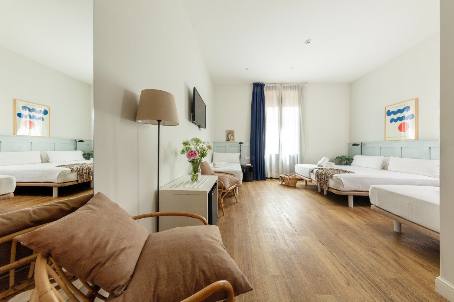 Una habitación de hotel luminosa y moderna con varias camas, suelos de madera clara, una silla de ratán.