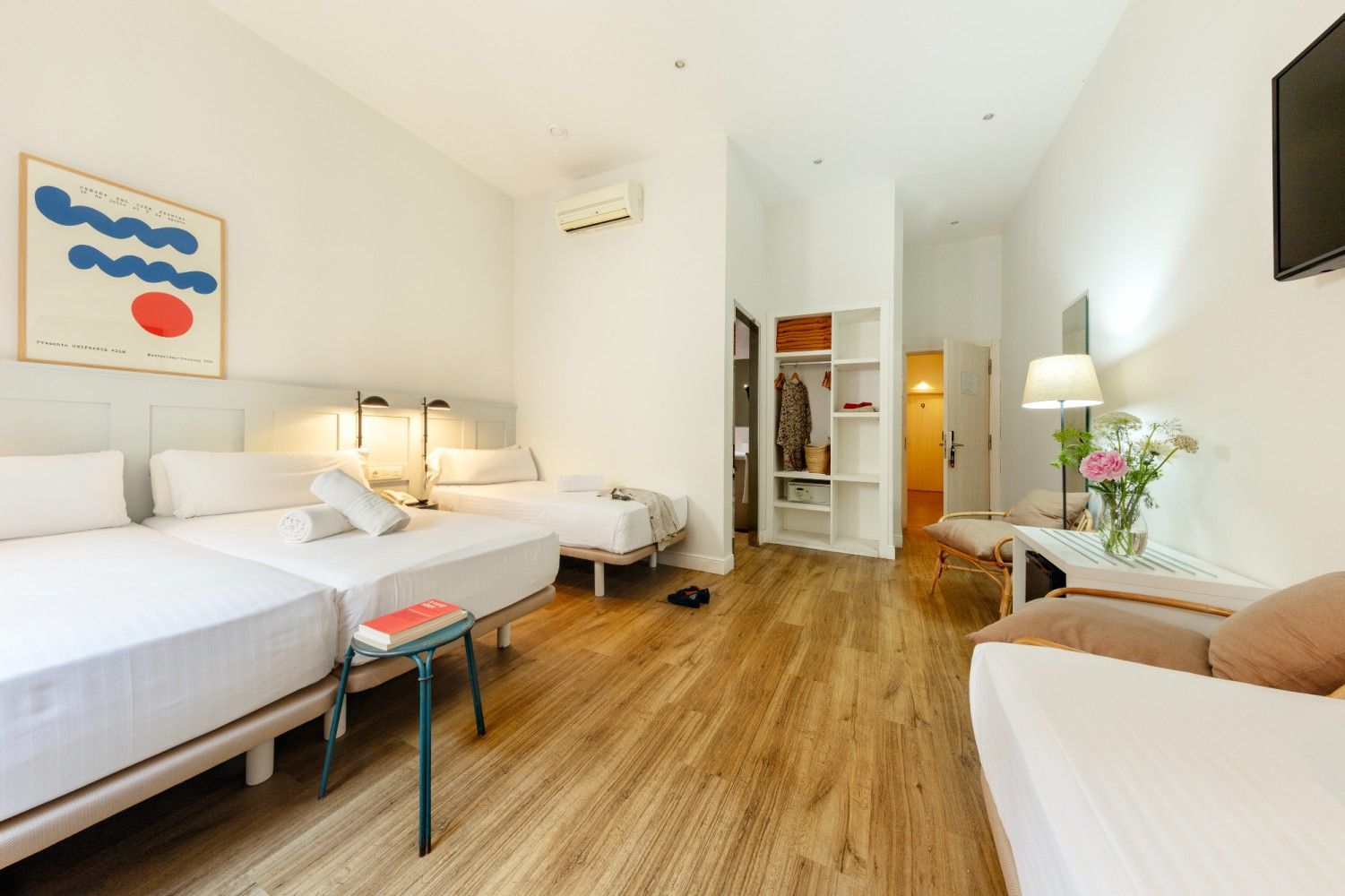 Una habitación de hotel con dos camas blancas, pisos de madera, un armario, un escritorio con lámpara y flores.