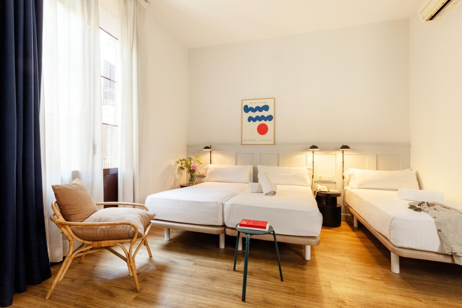 Una luminosa habitación de hotel con tres camas individuales, un sillón de ratán, suelos de madera.