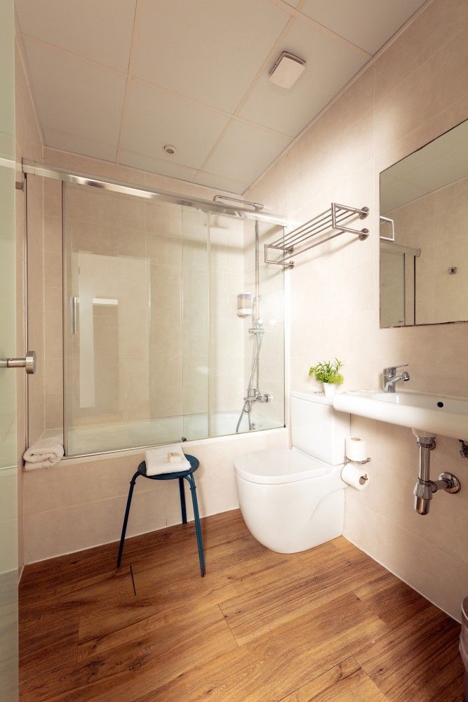 Un baño moderno y accesible en tonos claros con piso con apariencia de madera, ducha con mampara de vidrio, inodoro y lavabo.