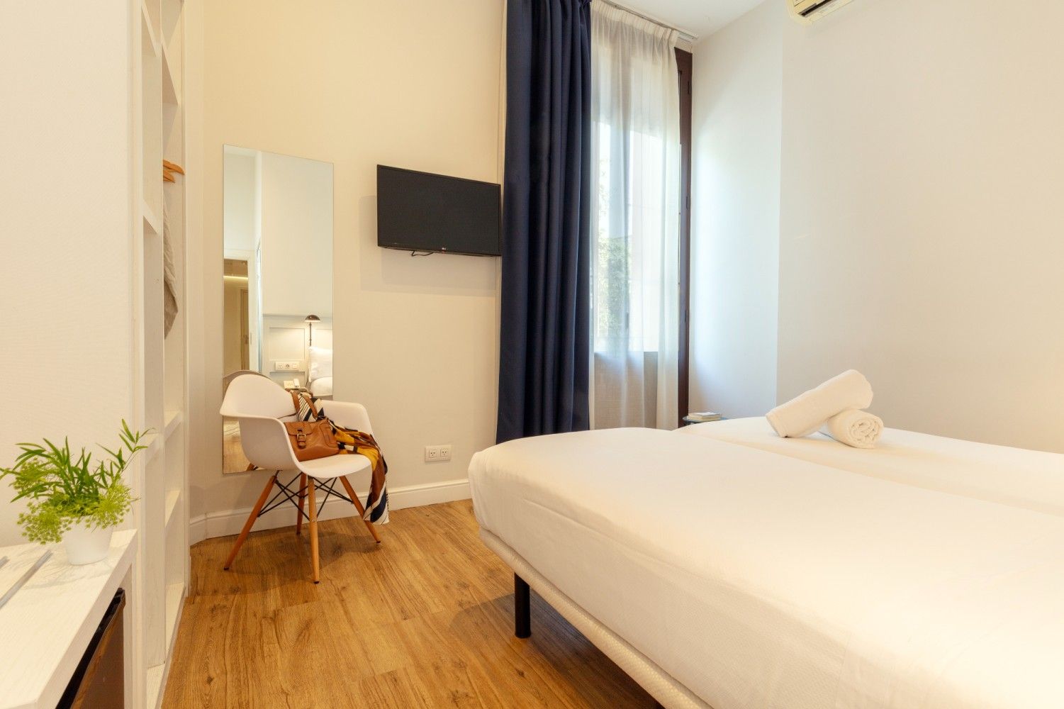 Una habitación de hotel luminosa y minimalista con una cama blanca, una pequeña silla blanca, un televisor.