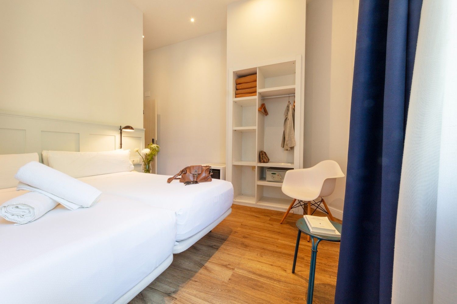 Un dormitorio con una cama grande con sábanas blancas, una estantería de madera, una silla blanca y una pequeña mesa.