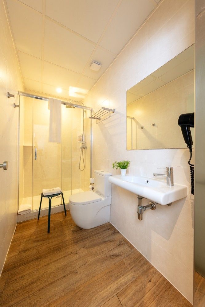 Un moderno baño de hotel con ducha de cristal, inodoro blanco, lavabo montado en la pared, espejo y suelo.