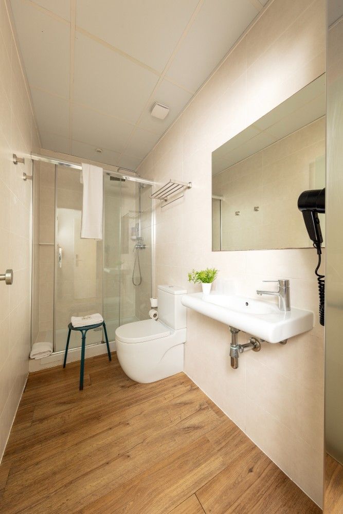 Un baño moderno con paredes claras, suelo de imitación madera, cabina de ducha de cristal, inodoro, lavabo y secador de pelo.