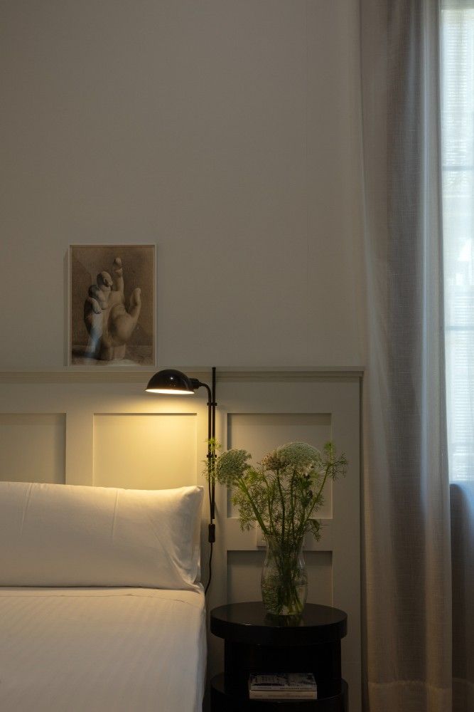 Un dormitorio de paredes blancas con una lámpara de pared encendida sobre la cabecera, una mesita de noche con flores.