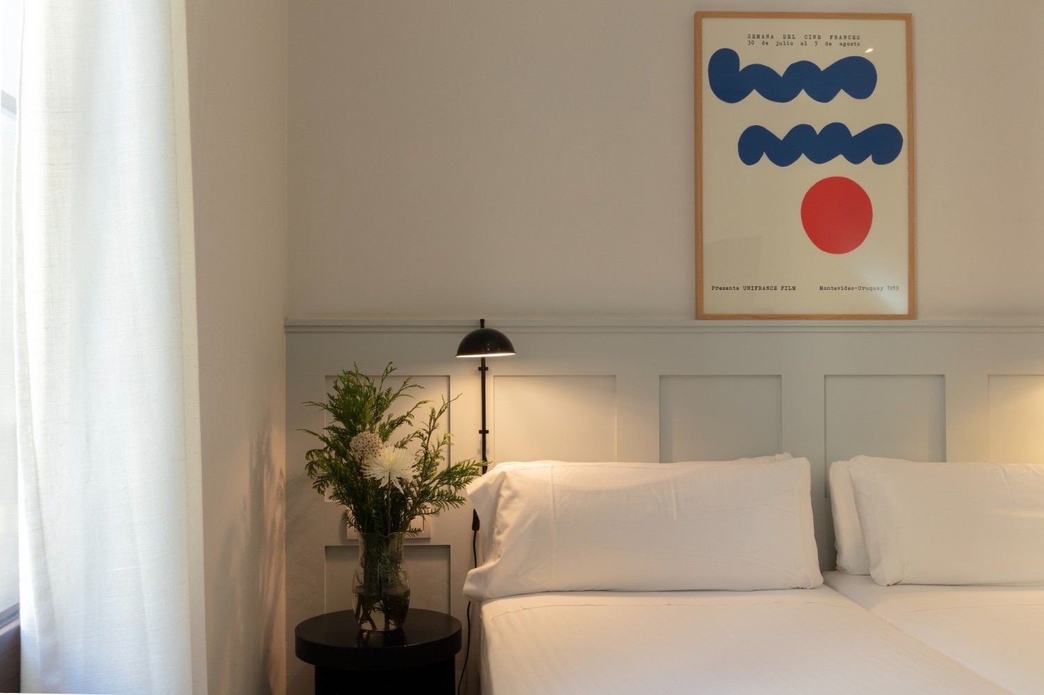 Un dormitorio luminoso con una cama, ropa de cama blanca, una mesita de noche negra con flores.