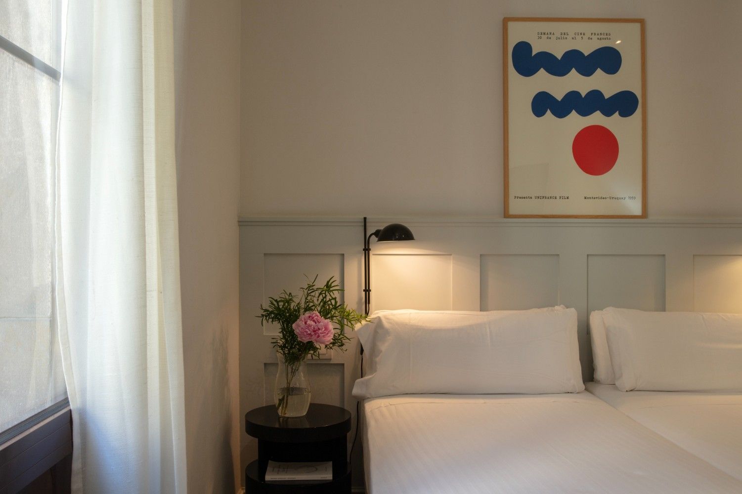 Un rincón de dormitorio con una cama, ropa de cama blanca, una mesa auxiliar negra con flores rosas.