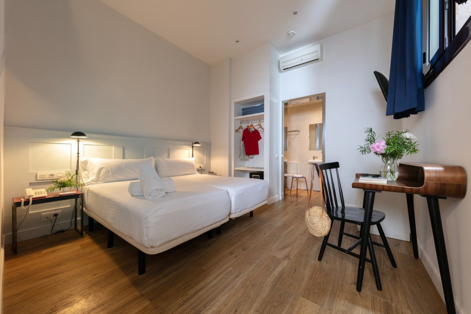 Una habitación de hotel con dos camas blancas, un escritorio de madera con una silla, un pequeño armario con ropa.