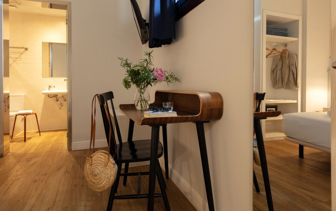 Un dormitorio moderno con un escritorio de madera y una silla negra, un jarrón de flores, un armario abierto.