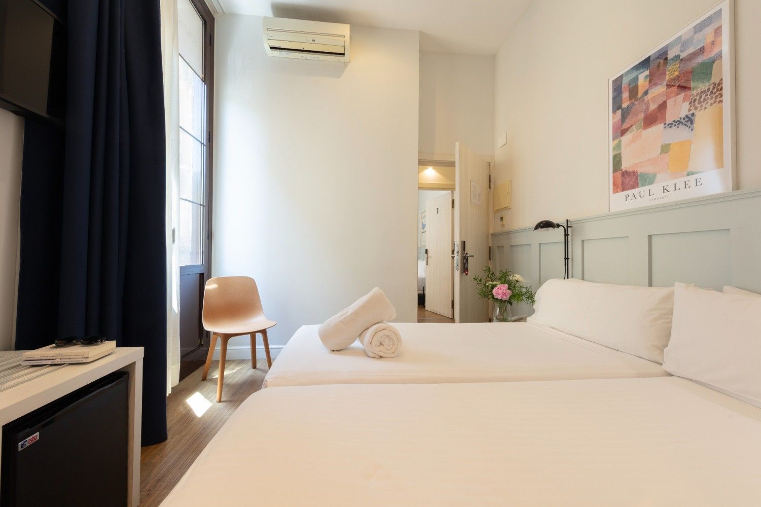 Una habitación de hotel luminosa y moderna con dos camas blancas, una silla junto a la ventana.