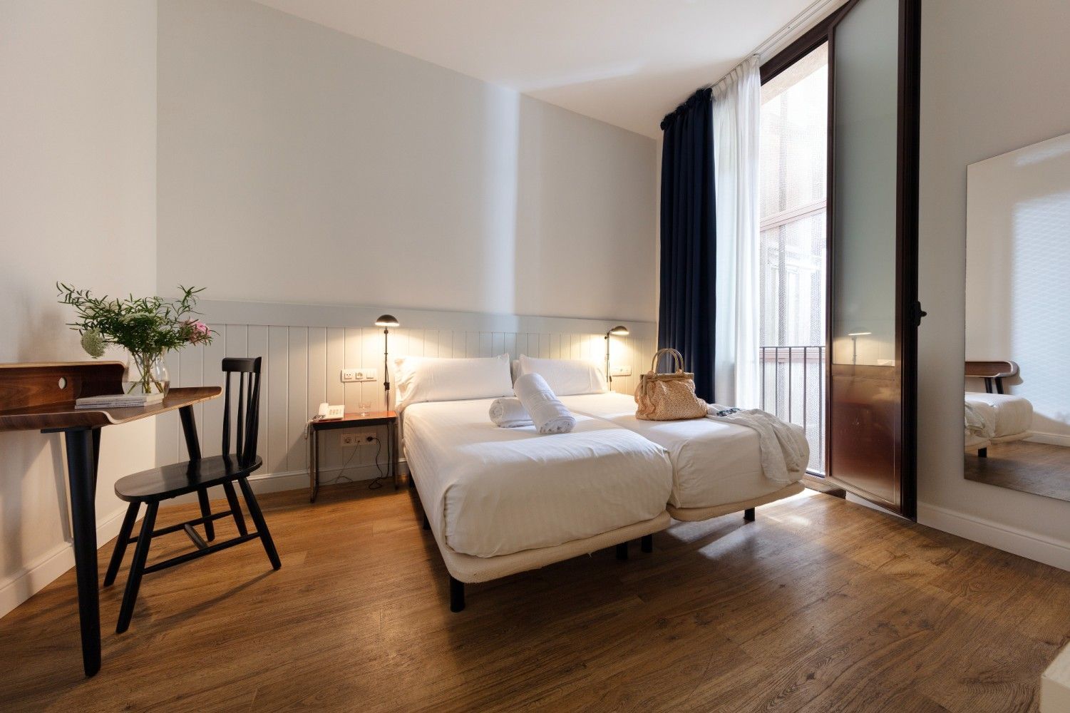 Una habitación de hotel luminosa y minimalista con dos camas individuales, un escritorio de madera con silla.