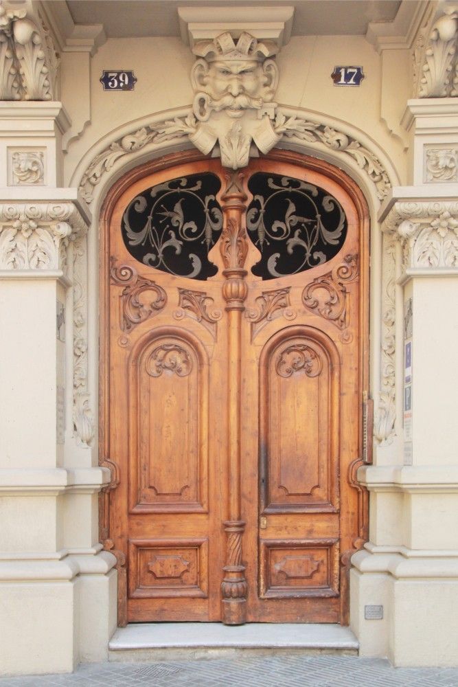 Puerta de madera adornada, de doble hoja, con una cara de piedra tallada en la parte superior.