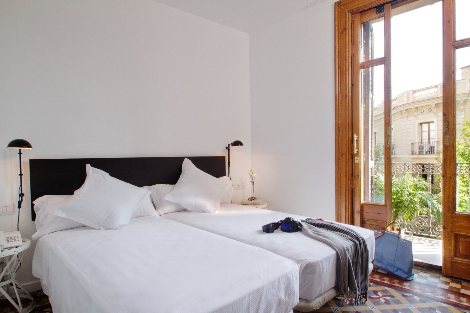 Un dormitorio luminoso con dos camas individuales con sábanas blancas, un cabecero oscuro y una puerta de balcón.