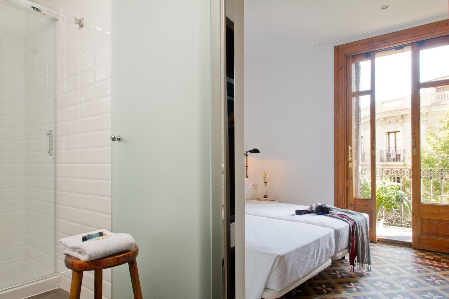 Una vista a través de una puerta que muestra un baño con ducha blanca y taburete junto a un dormitorio con dos camas.