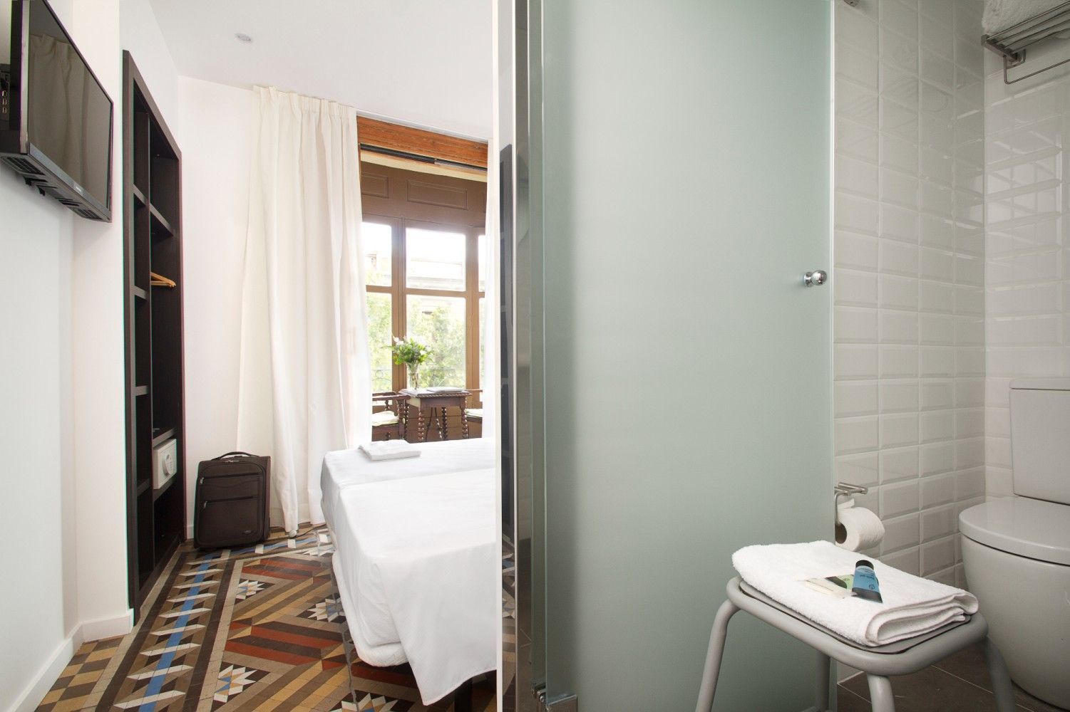 Vista de una habitación de hotel con piso estampado, cama blanca y un baño adyacente con ducha y WC.