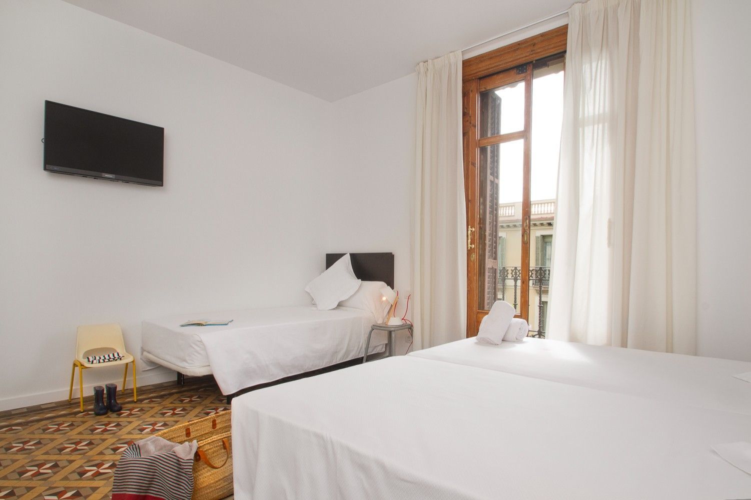 Una habitación de hotel con dos camas blancas, un televisor montado en la pared, una silla y un balcón con cortinas blancas.