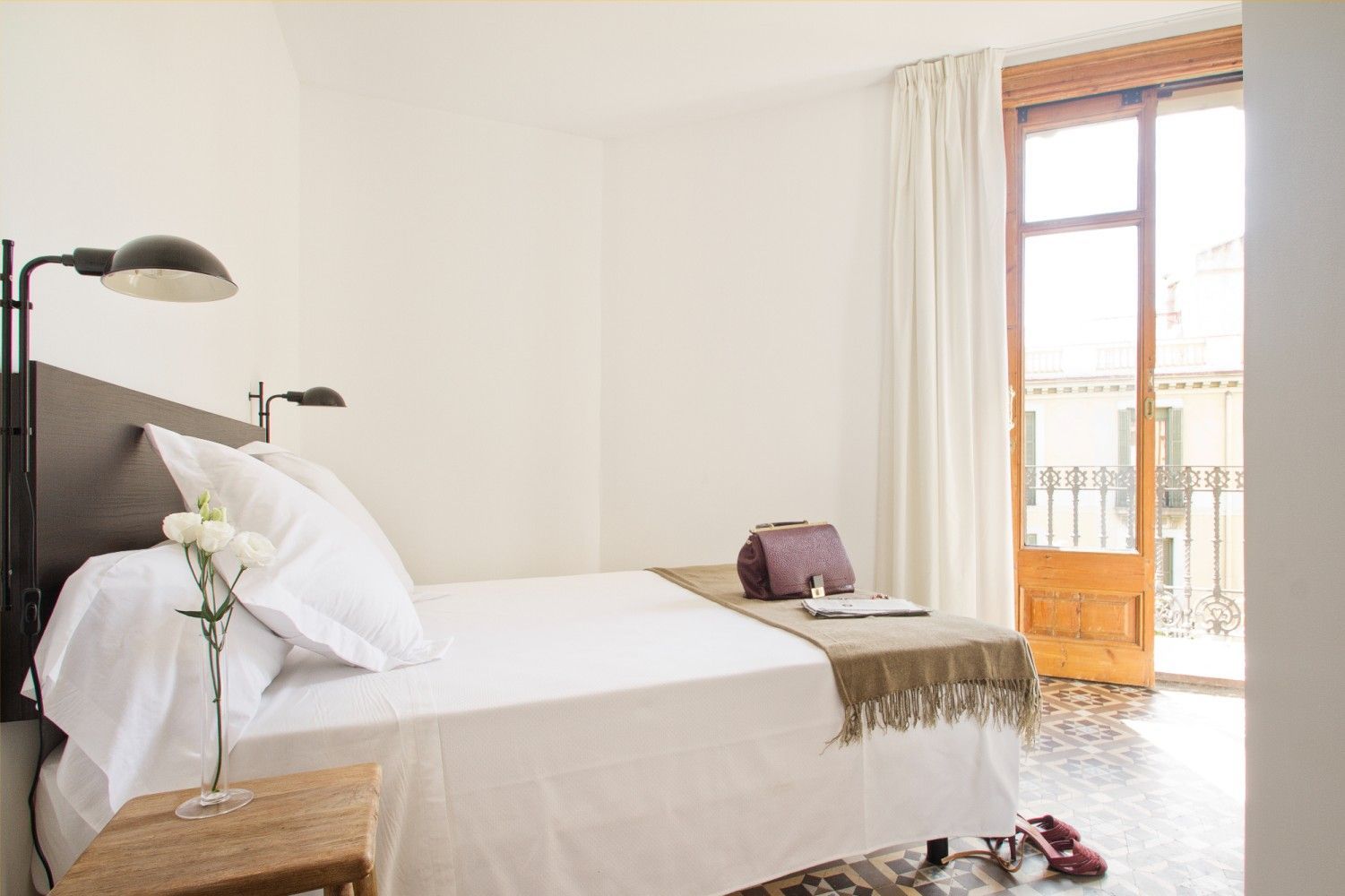 Un dormitorio luminoso con una cama blanca, dos lámparas de pared y una puerta de balcón.