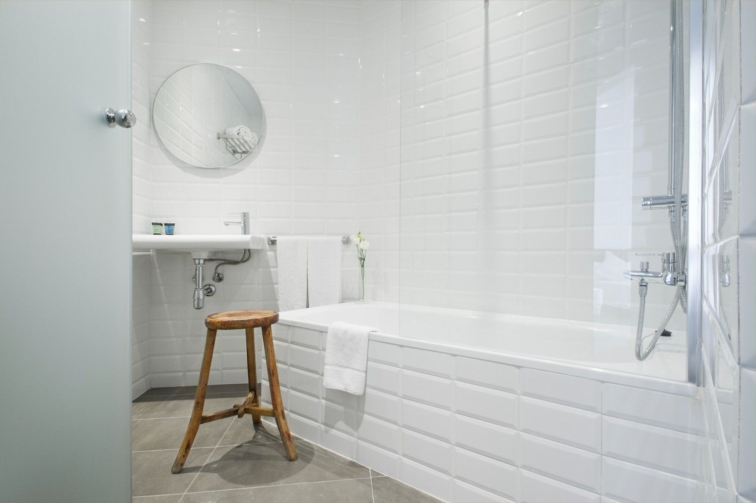 Un baño moderno y minimalista con paredes de azulejos blancos, bañera con ducha, espejo circular y un taburete de madera.