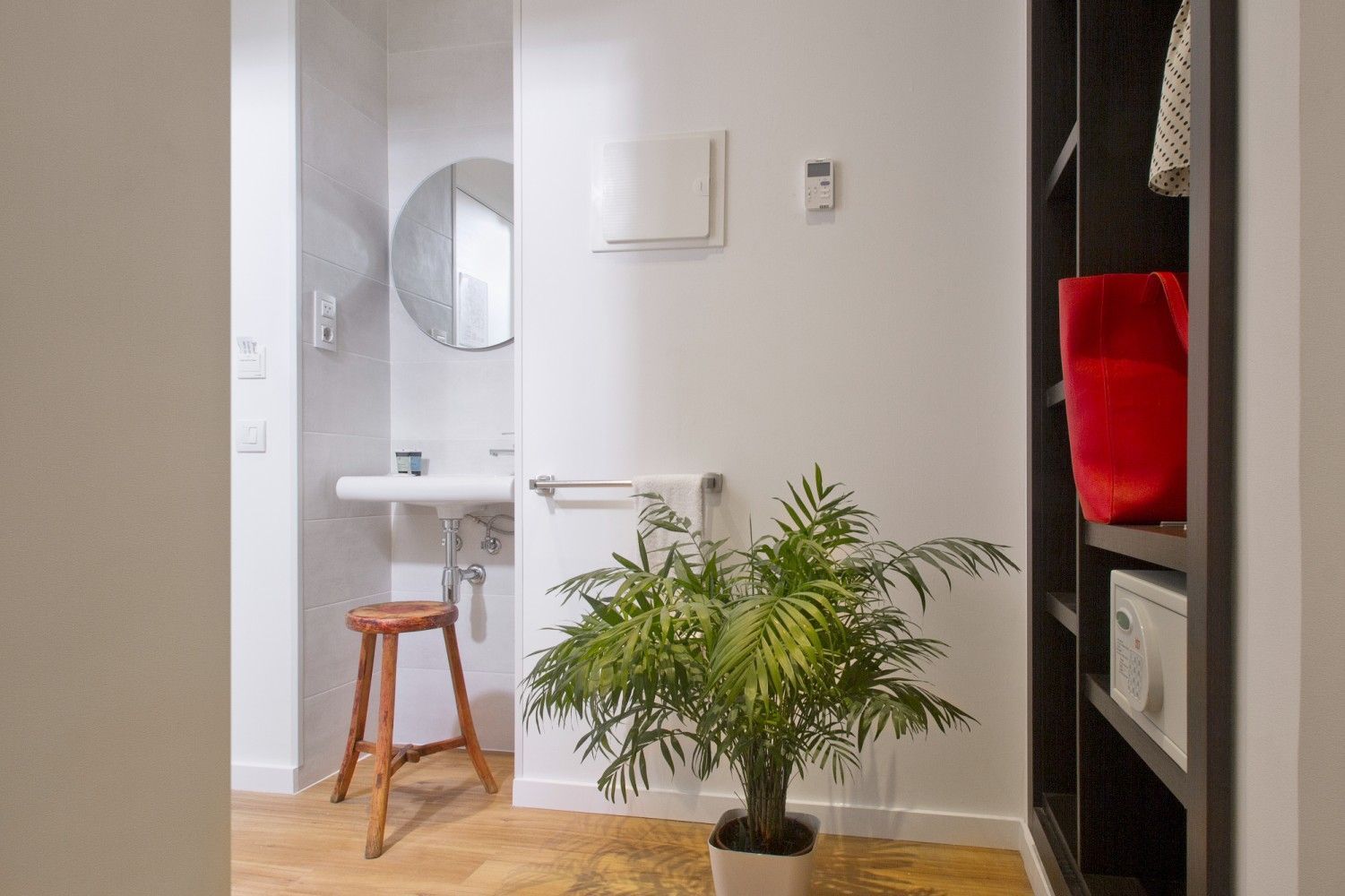 Una habitación limpia y minimalista con un taburete de madera, un lavabo blanco, un espejo, una planta verde en maceta.
