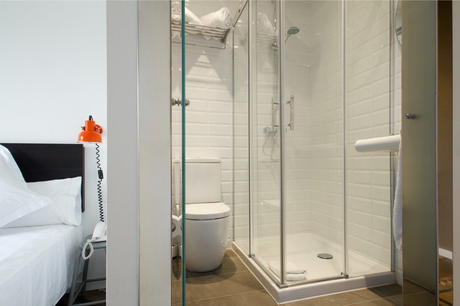 Vista de una habitación de hotel que da a un pequeño baño con azulejos blancos y una ducha con mampara de vidrio.