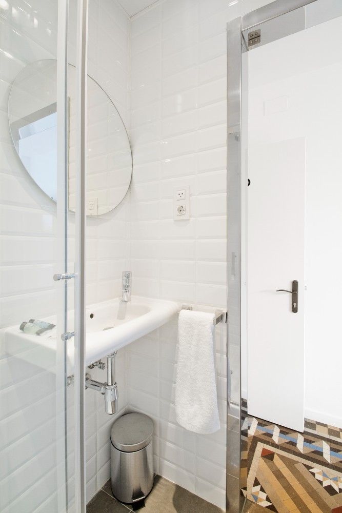 Un baño pequeño y luminoso con paredes de azulejos blancos, un espejo redondo, un lavabo con pedesta.