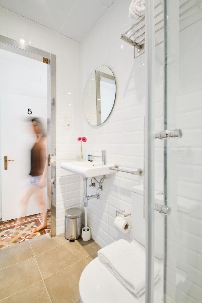 Un baño luminoso y alicatado con lavabo, inodoro, ducha y una figura borrosa que pasa por una puerta abierta.