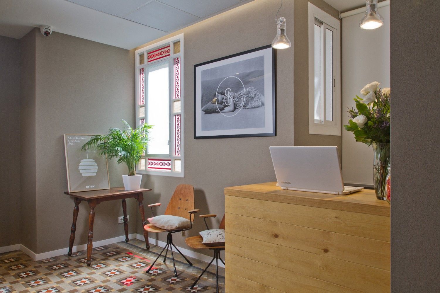 Un área de recepción con un escritorio de madera, una computadora portátil, dos sillas, una mesa pequeña con una planta.