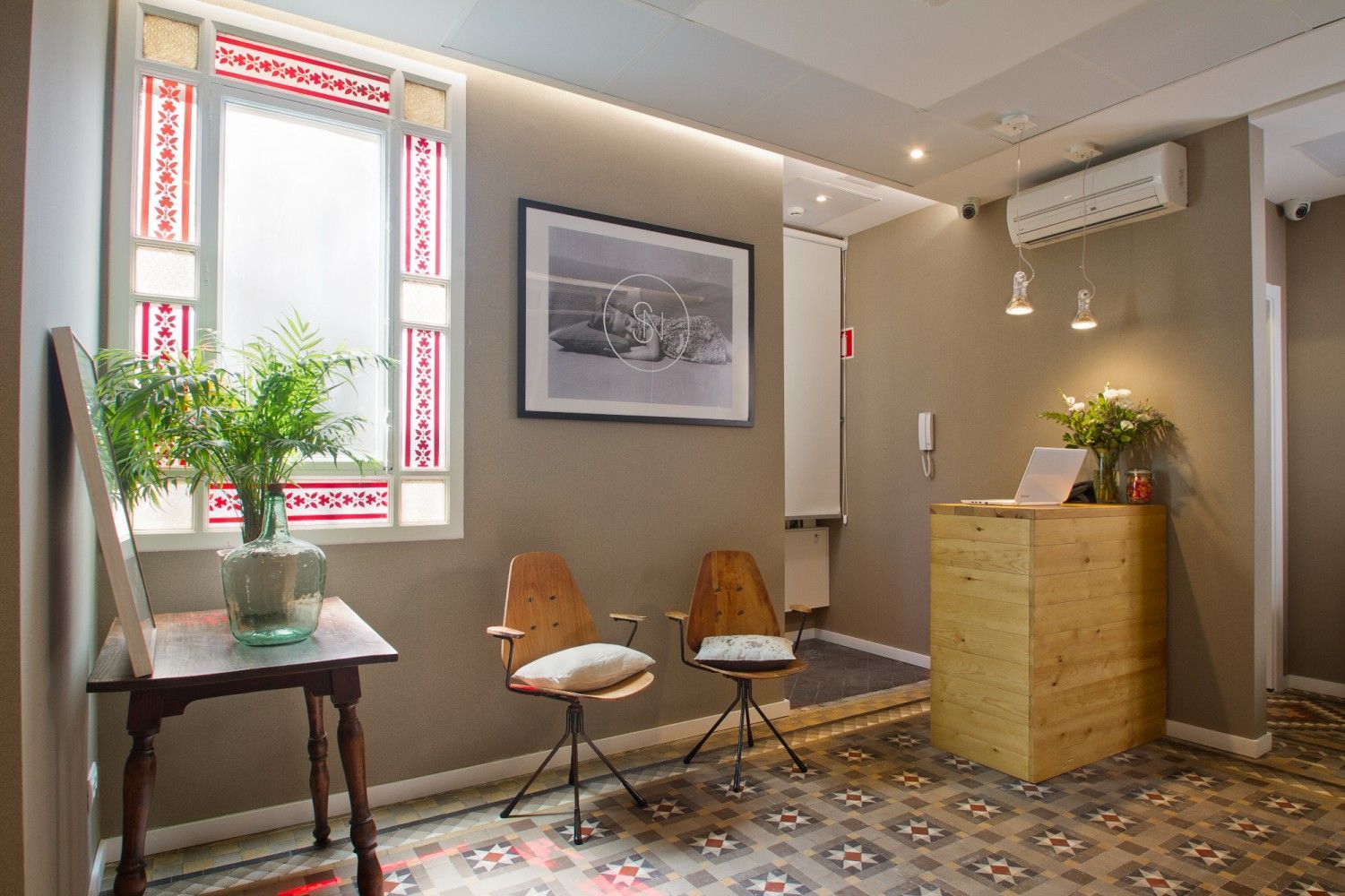 Un área de recepción con dos sillas de madera, una mesa auxiliar con una planta, una impresión en blanco .