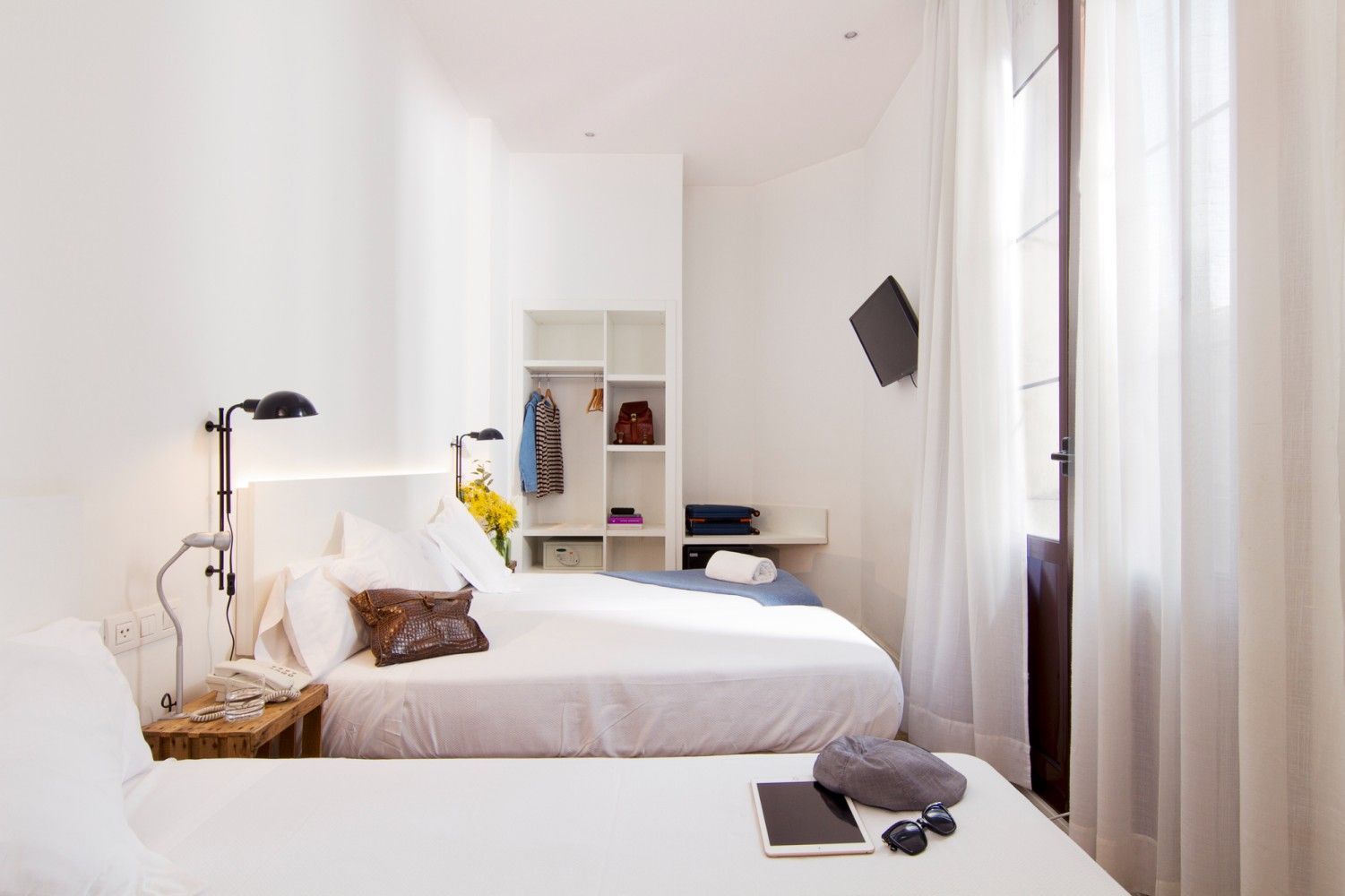 Una moderna habitación de hotel con dos camas blancas, un televisor montado en la pared, una estantería abierta.