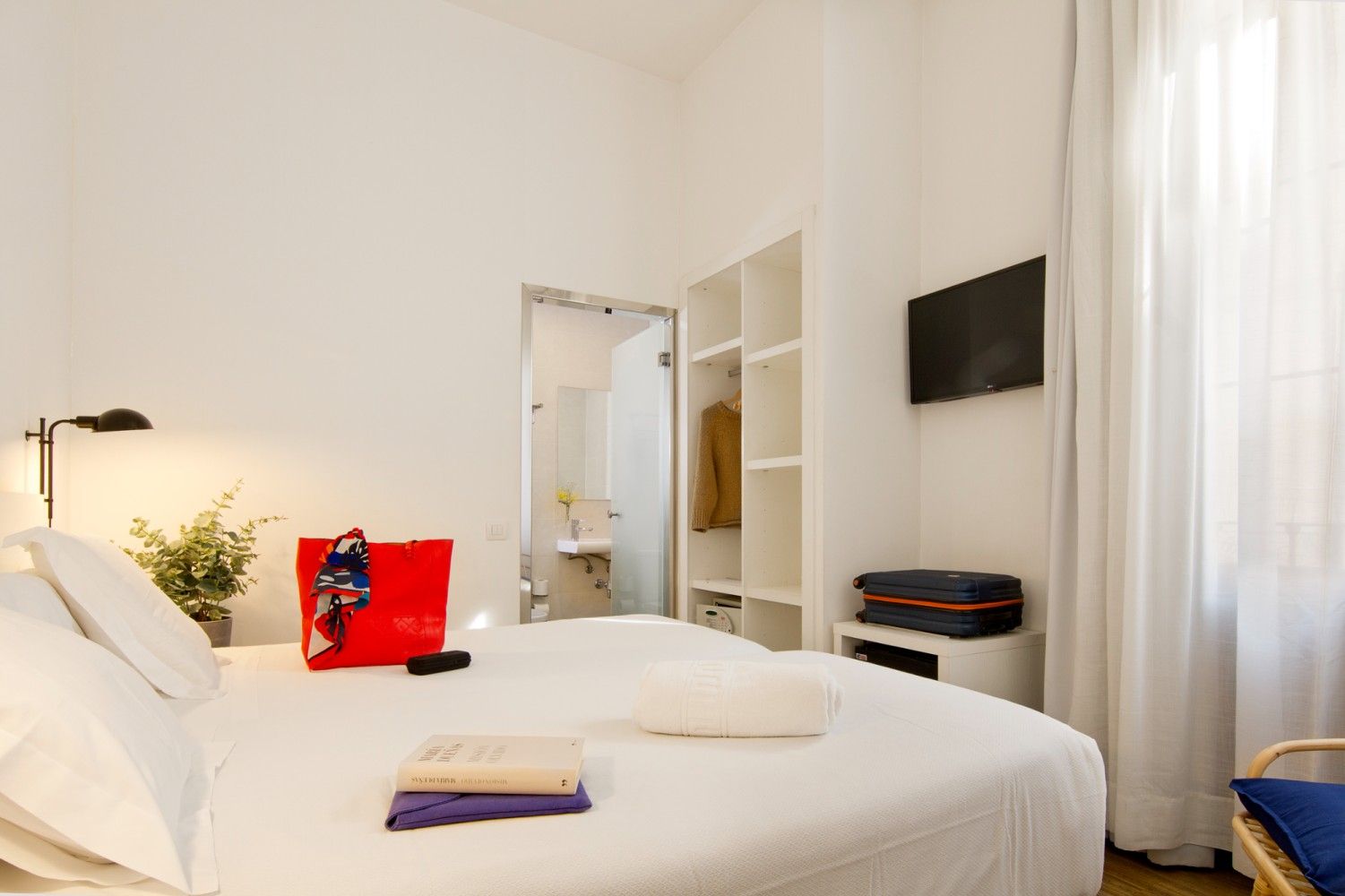 Una habitación de hotel blanca y luminosa con una cama cuidadosamente hecha, un bolso rojo, un televisor, un armario.