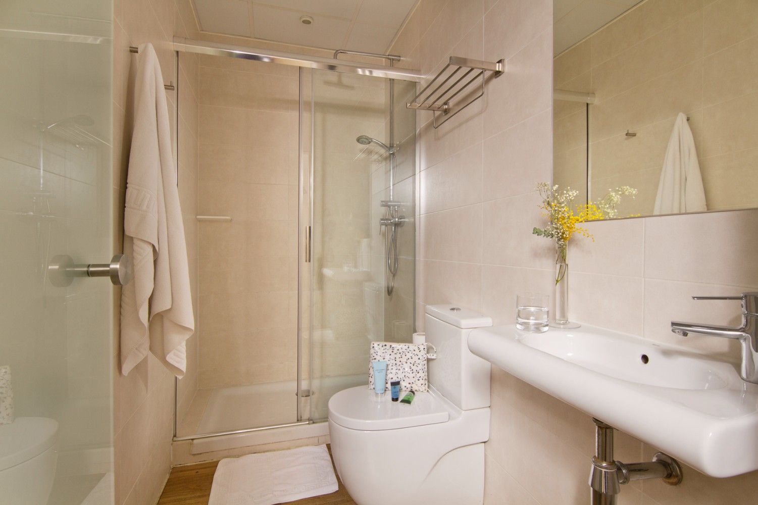 Un baño beige con una ducha con mampara de cristal, un inodoro montado en la pared, un lavabo blanco y una toalla colgada.