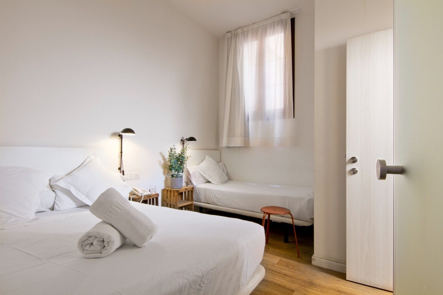 Un dormitorio de hotel con una cama grande en primer plano, una cama individual junto a la ventana y una puerta blanca.