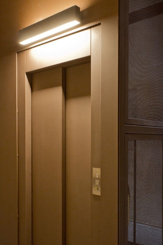 Una puerta de ascensor cerrada con un botón de llamada a la derecha y una lámpara iluminada sobre el marco en un pasillo.