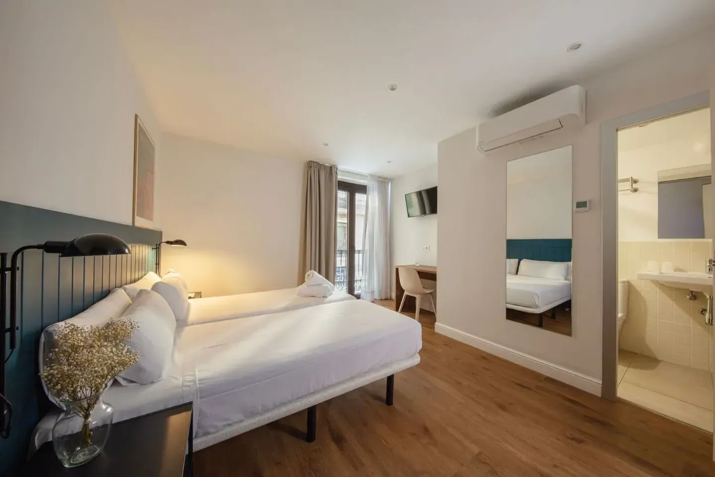 Una moderna habitación de hotel con dos camas individuales blancas, pisos de madera, un espejo y un baño adyacente.