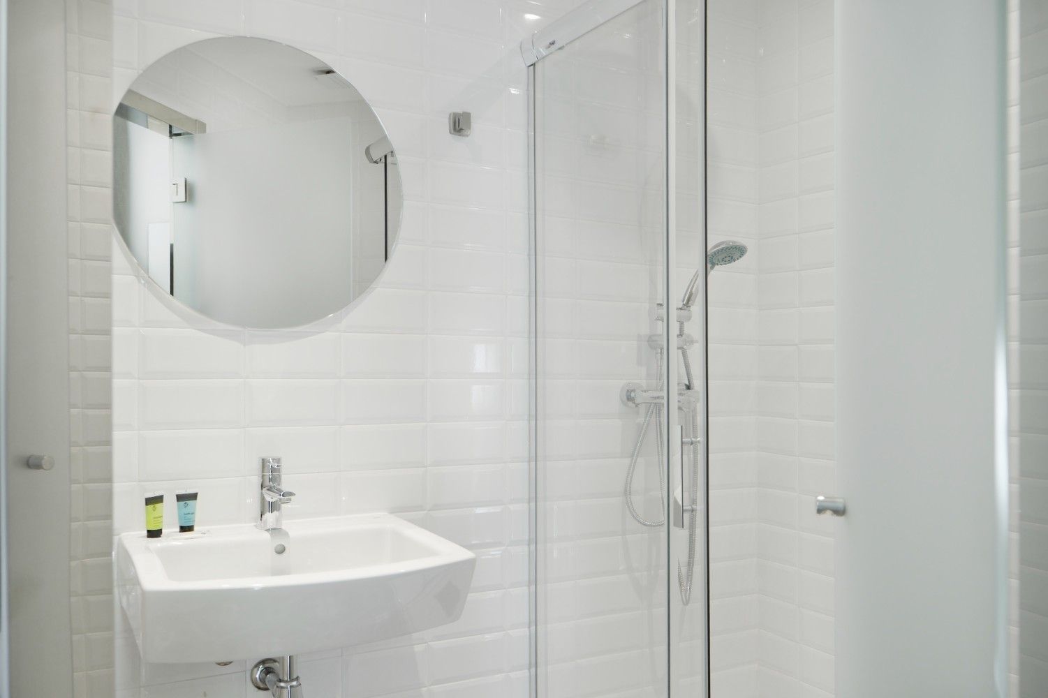 Un baño blanco con un espejo redondo sobre un lavabo montado en la pared, al lado de una ducha con mampara de cristal.