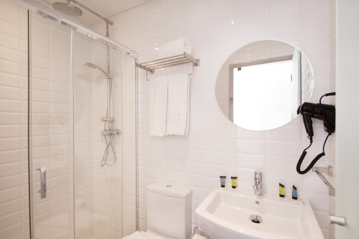Un baño blanco moderno y luminoso que cuenta con una cabina de ducha de cristal, inodoro, lavabo, secador de pelo.