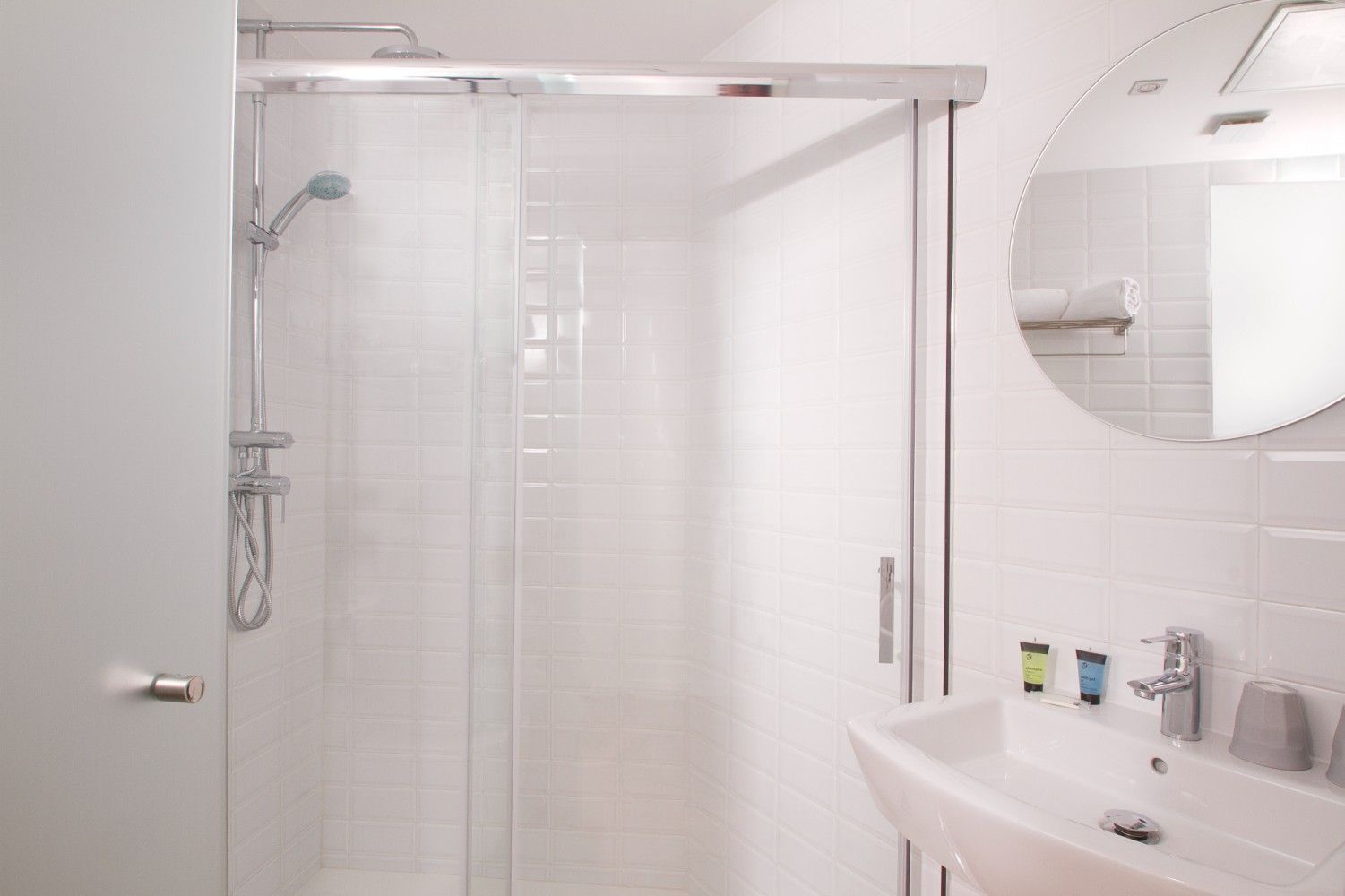 Un baño moderno con ducha de azulejos blancos con puerta de cristal, lavabo montado en la pared y espejo circular.