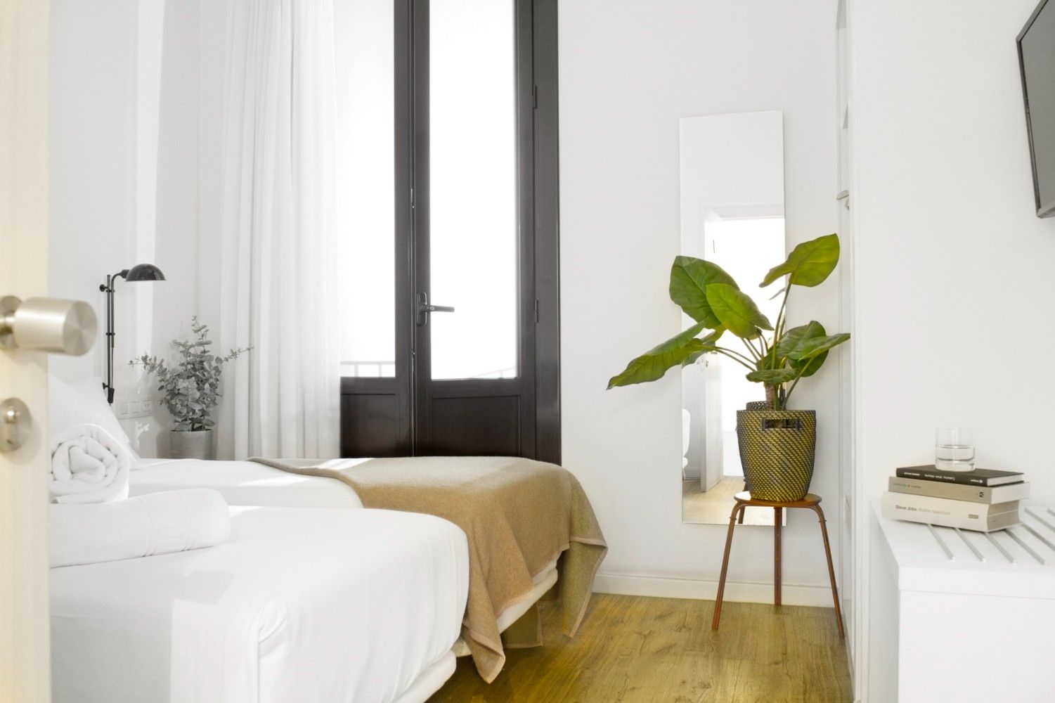 Un dormitorio de hotel minimalista y blanco con dos camas individuales, un espejo de piso alto, una planta en maceta.