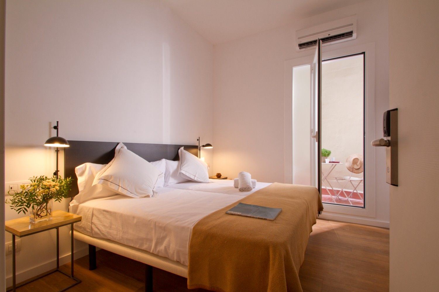 Una moderna habitación de hotel con una cama cuidadosamente hecha, una pequeña mesa auxiliar con una planta y una puerta.