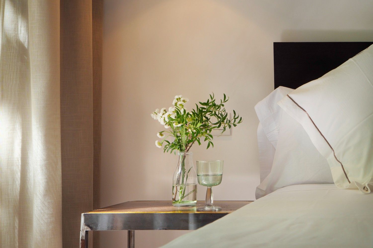 Un jarrón de vidrio con flores blancas y un vaso de agua se encuentran sobre una mesa de noche de metal.