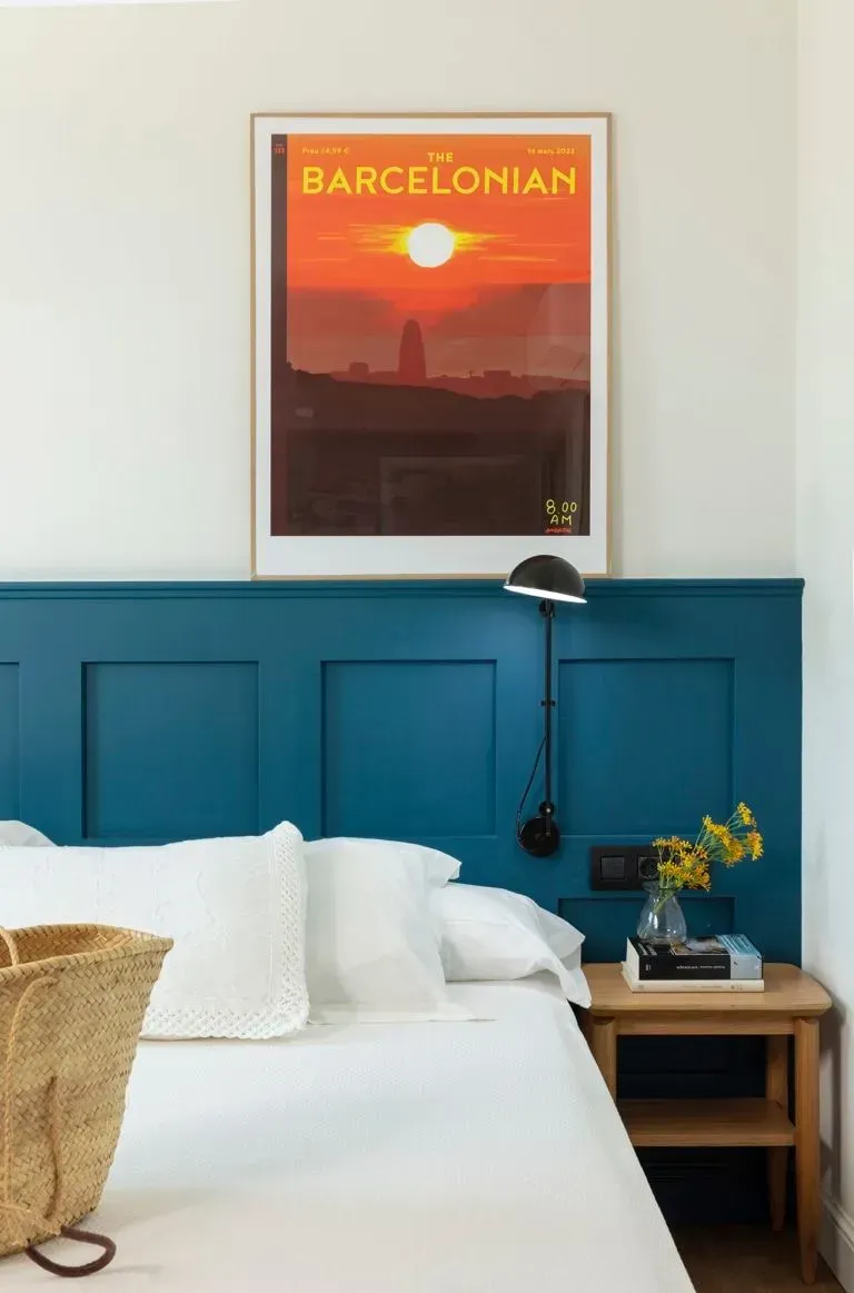 Detrás de una cama blanca hay un cabecero azul con un póster de una puesta de sol 