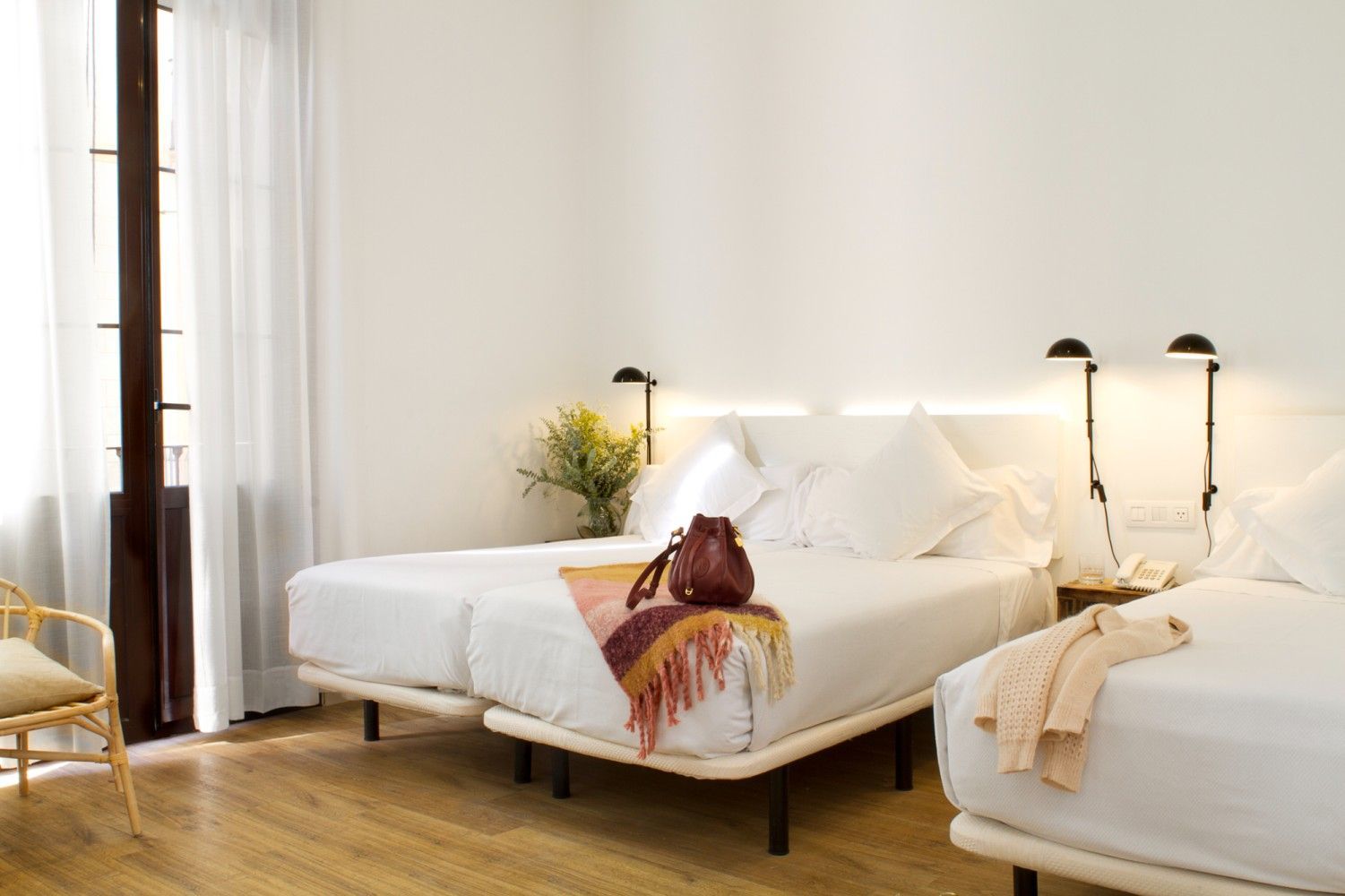 Una luminosa habitación de hotel cuenta con dos camas individuales con sábanas blancas, suelo de madera, cortinas