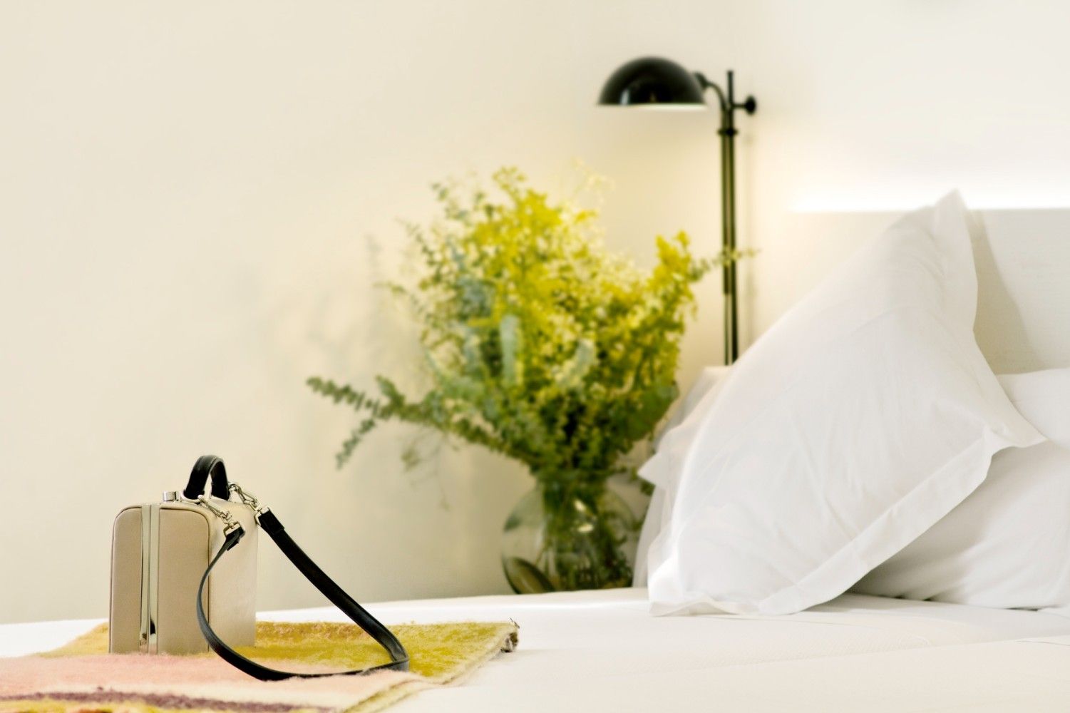 Un pequeño bolso reposa sobre una cama junto a un jarrón de cristal con flores amarillas y una lámpara de noche.